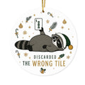 Round acrylic ornament with a tired raccoon in a green Santa hat holding up a mahjong tile and the words Discarded the Wrong Tile